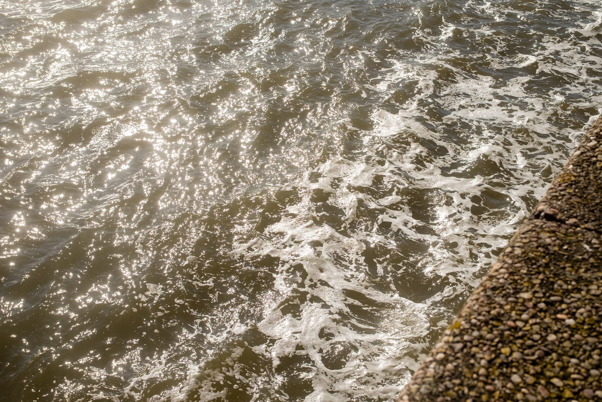 Close-up view of sunlit water with small waves and a textured stone wall in the corner.