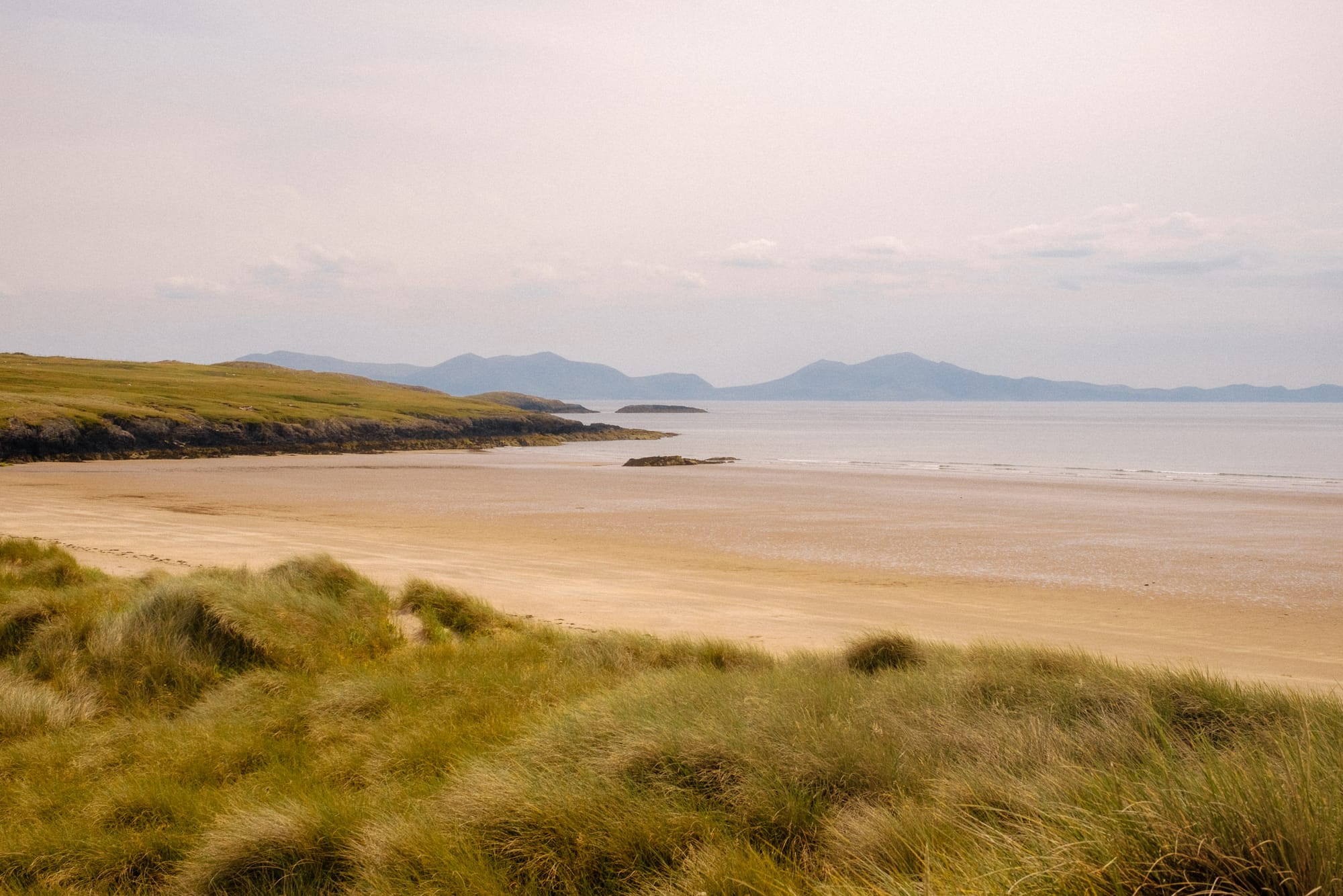 A tranquil coastal scene featuring a sandy beach lined with gentle dunes covered in tall grass. In the background, rolling hills and mountains stretch towards the horizon under a soft, pastel sky. The calm sea reflects the muted colors of the surroundings, creating a peaceful and serene atmosphere.
