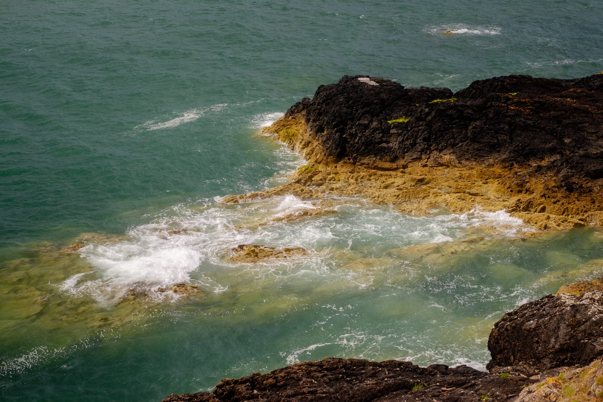 A coastal scene featuring rocky shoreline with dark, rugged rocks contrasting against the turquoise water. Waves gently break against the rocks, creating white frothy edges, while the clear water reveals some submerged rocks and a sandy bottom. The overall atmosphere is serene, with the ocean's movement creating a sense of calm.