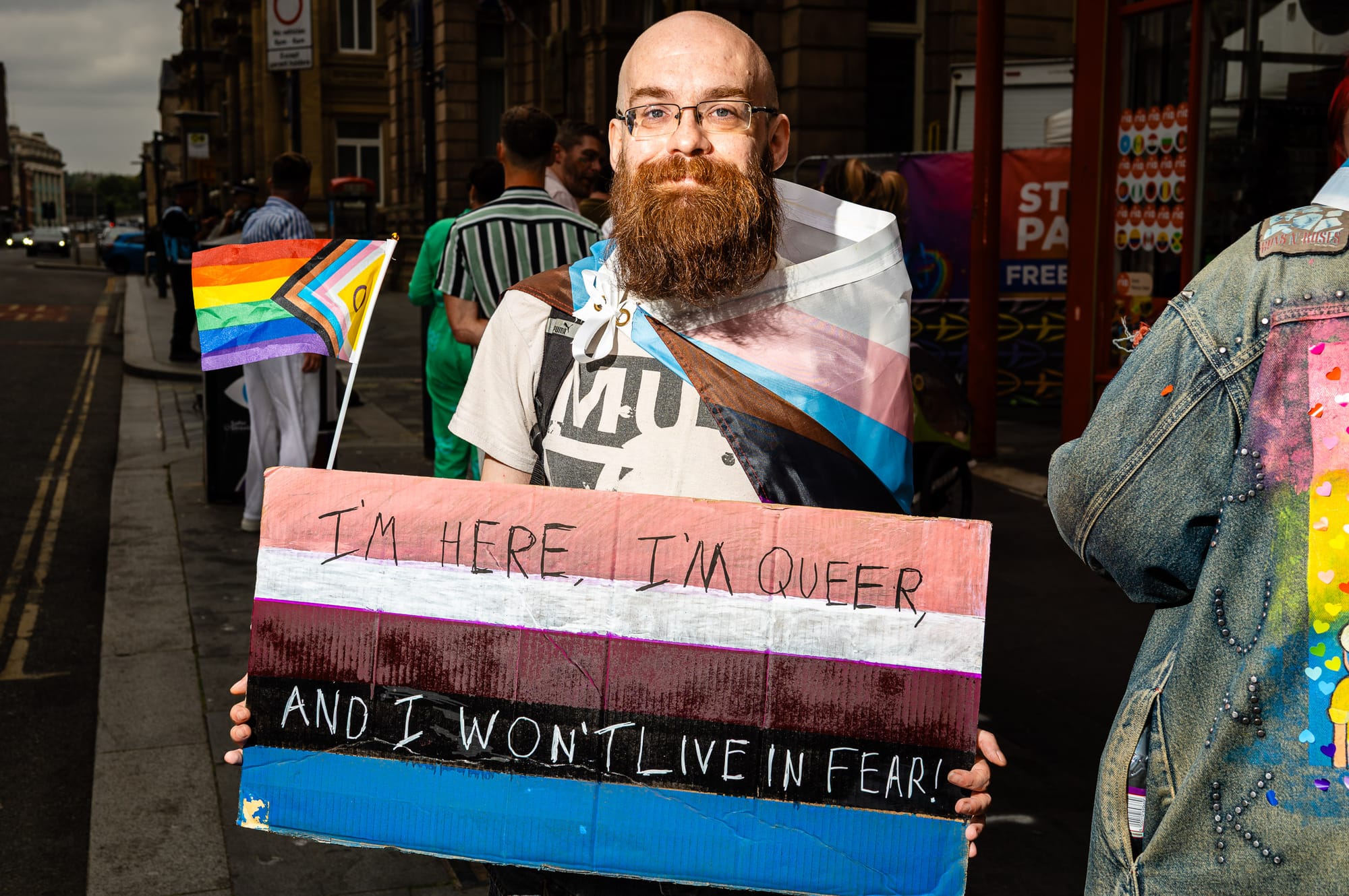 A bearded person wears a trans flag and holds up a sign saying "I'm here, I'm queer, and I won't live in fear."