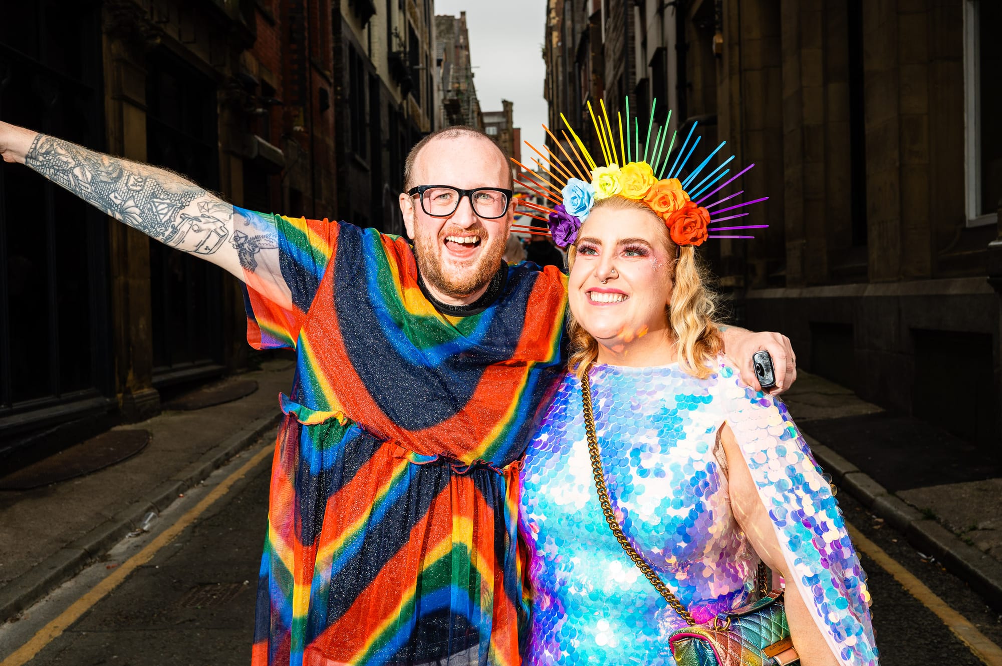 Two people at Liverpool Pride. One person with a beard wears a rainbow dress. The other wears a blue and pink sequin dress with a rainbow crown.