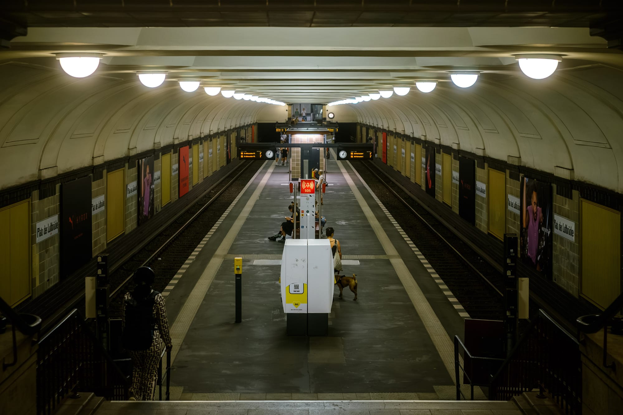 Interior view of a subway station in Berlin, Germany, featuring people, a dog, and advertising posters.