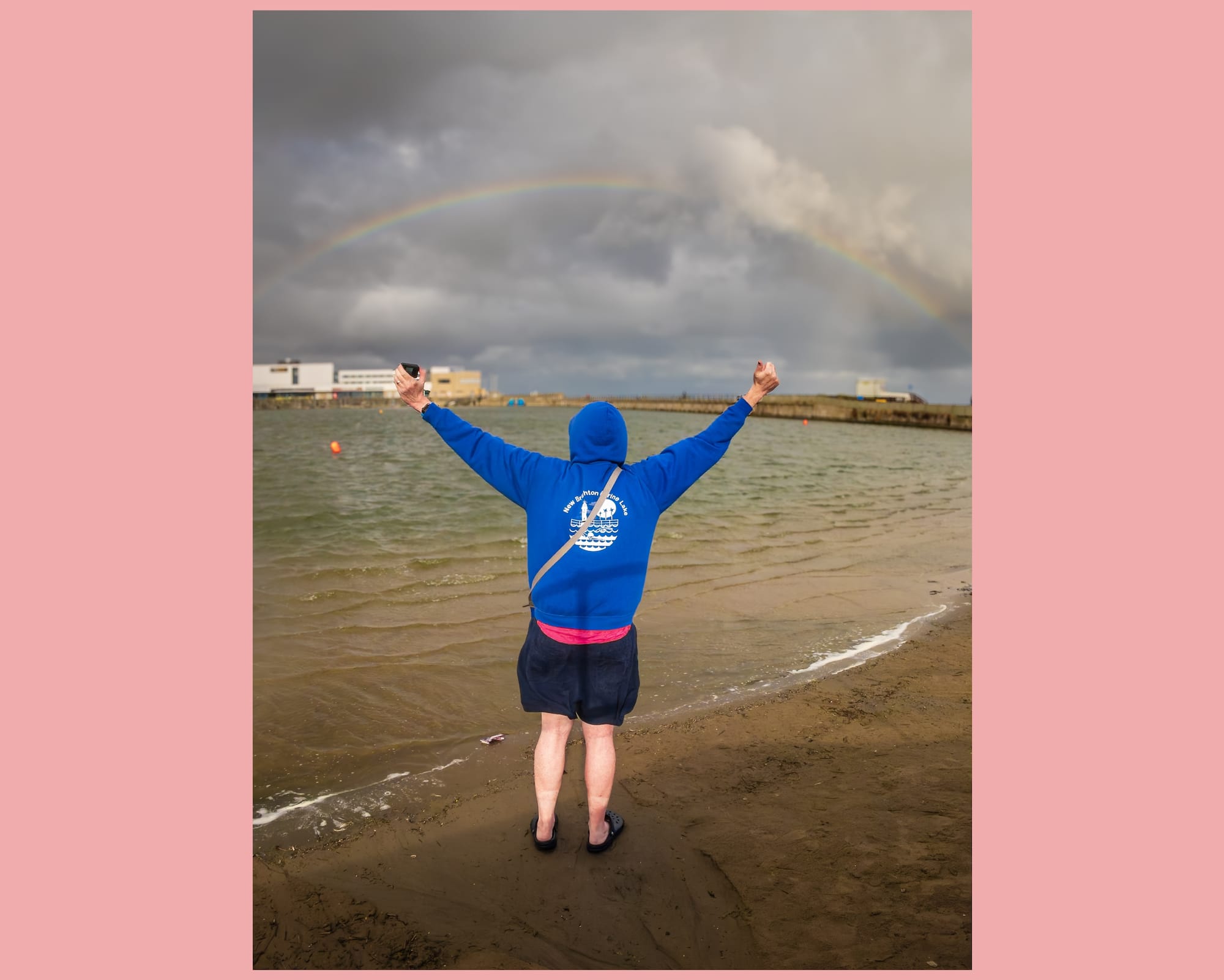 A person in a blue hoodie holds up their arms in celebration at the edge of a lake while a rainbow goes across the sky.