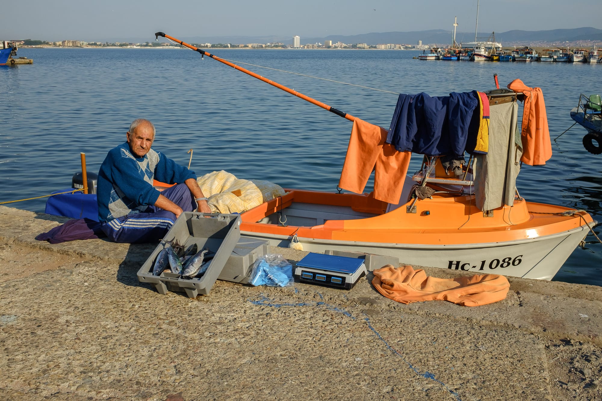 An old man in blue clothes sits by his orange boat selling freshly caught fish.