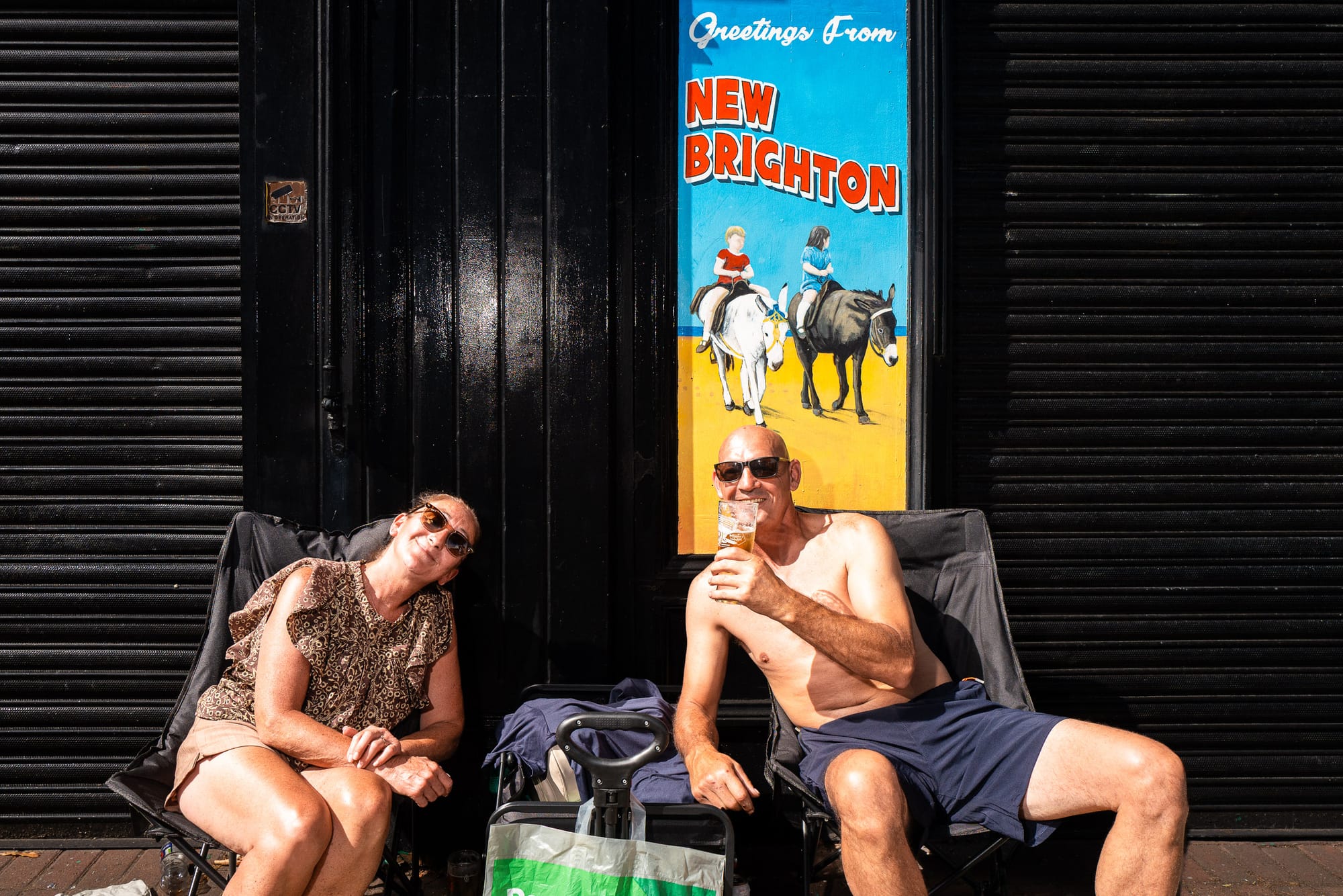 Two people in deck chairs outside a closed shop. A sign says "Greetings from New Brighton".