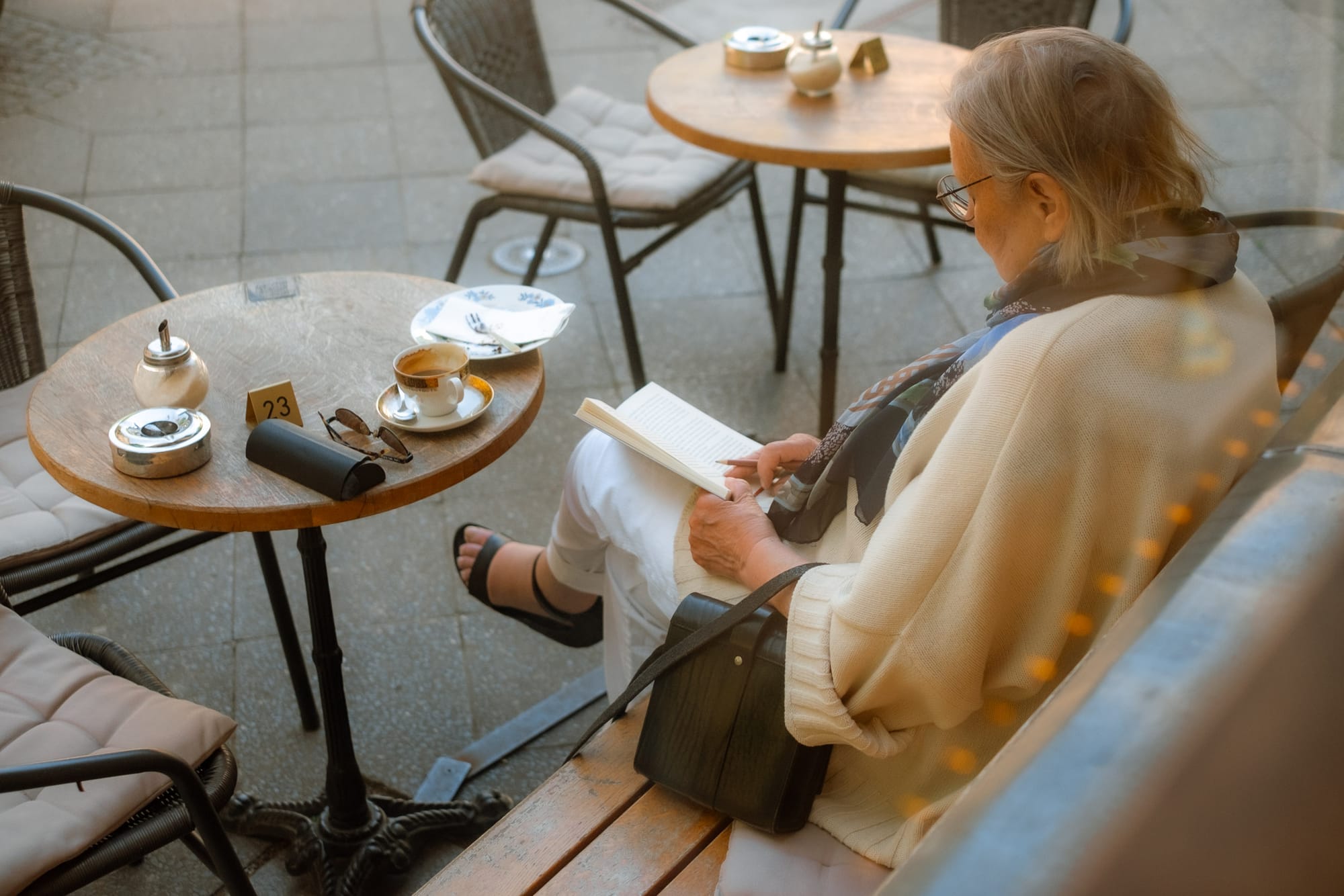 A woman seated at an outdoor café table reads a book while sipping coffee. She wears a light sweater and a scarf, with her hair pulled back. The setting features a few small tables and chairs, with a serene atmosphere. Sunlight filters through, creating a warm ambiance.