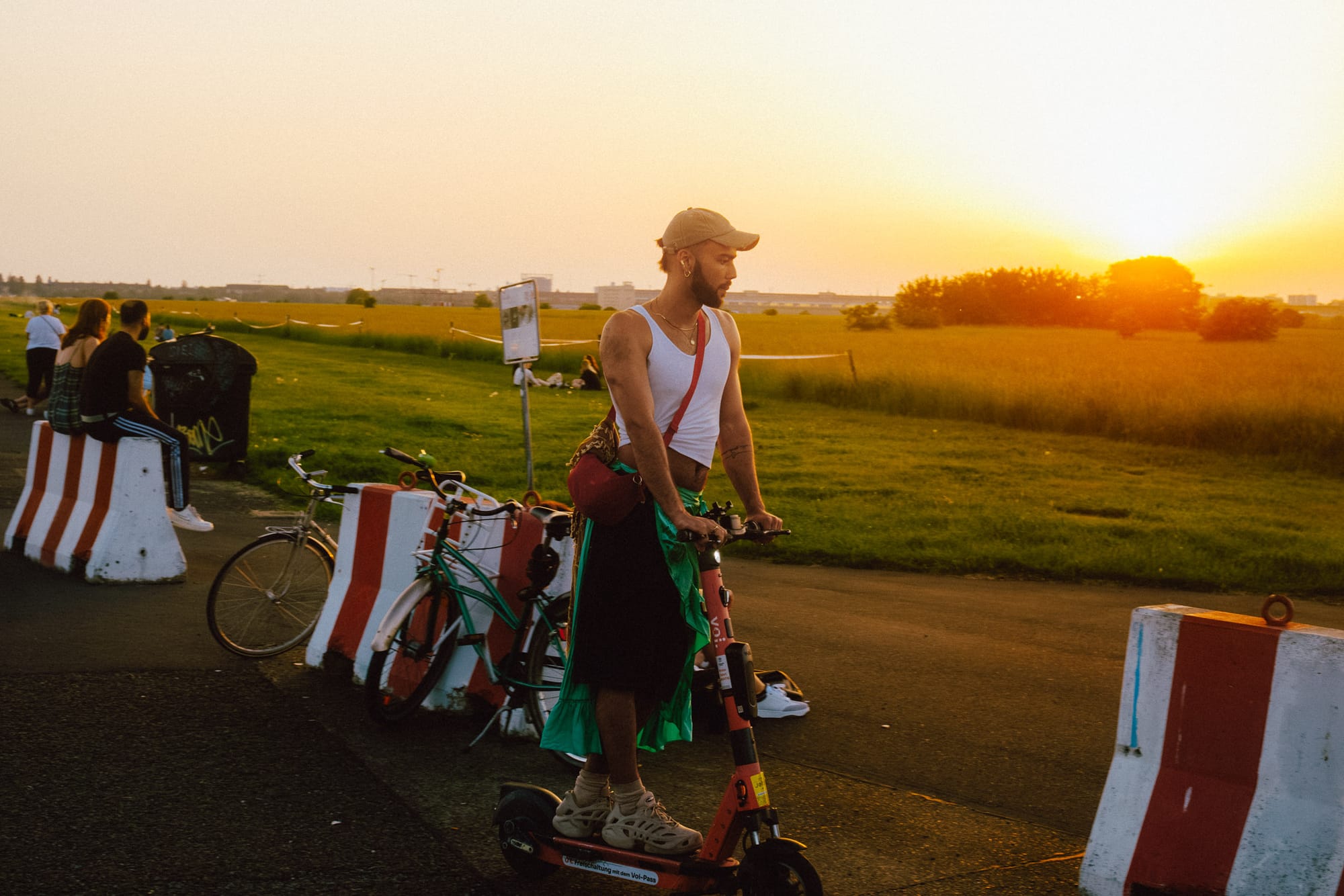 A person rides a scooter along a path during sunset, wearing a white tank top and a cap. In the background, a golden sun dips below the horizon, illuminating the landscape of green fields. Nearby, other individuals are seated on barriers, and bicycles are parked against white and red striped blocks. The scene conveys a relaxed, vibrant atmosphere typical of an outdoor setting during the evening.