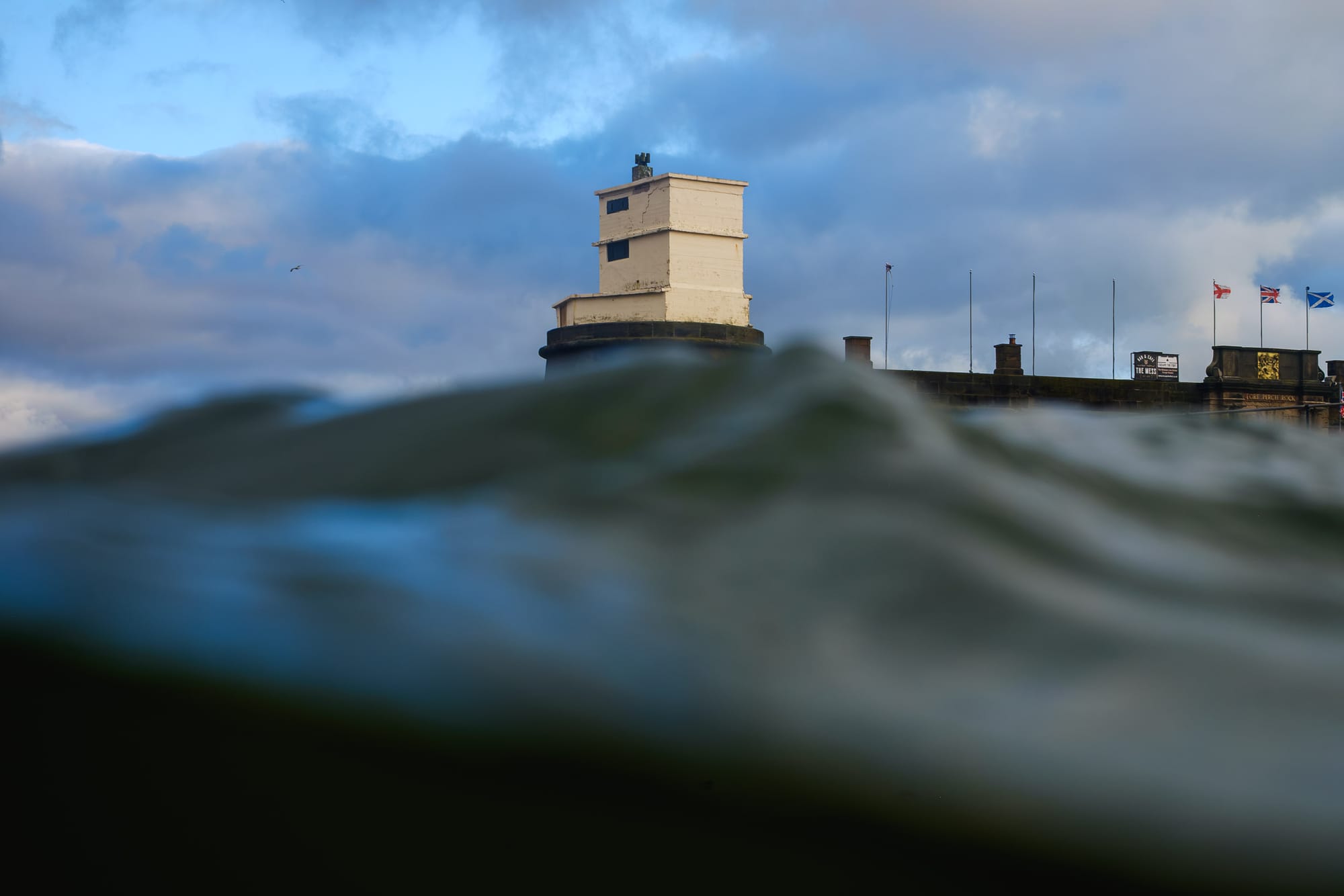 A view from the water’s surface, capturing gentle waves rolling toward a historic building on the shore. The structure, with a white facade and multiple flags flying on its roof, stands against a backdrop of a cloudy sky that hints at a changing weather. The scene conveys a sense of tranquility and connection to the marine environment.