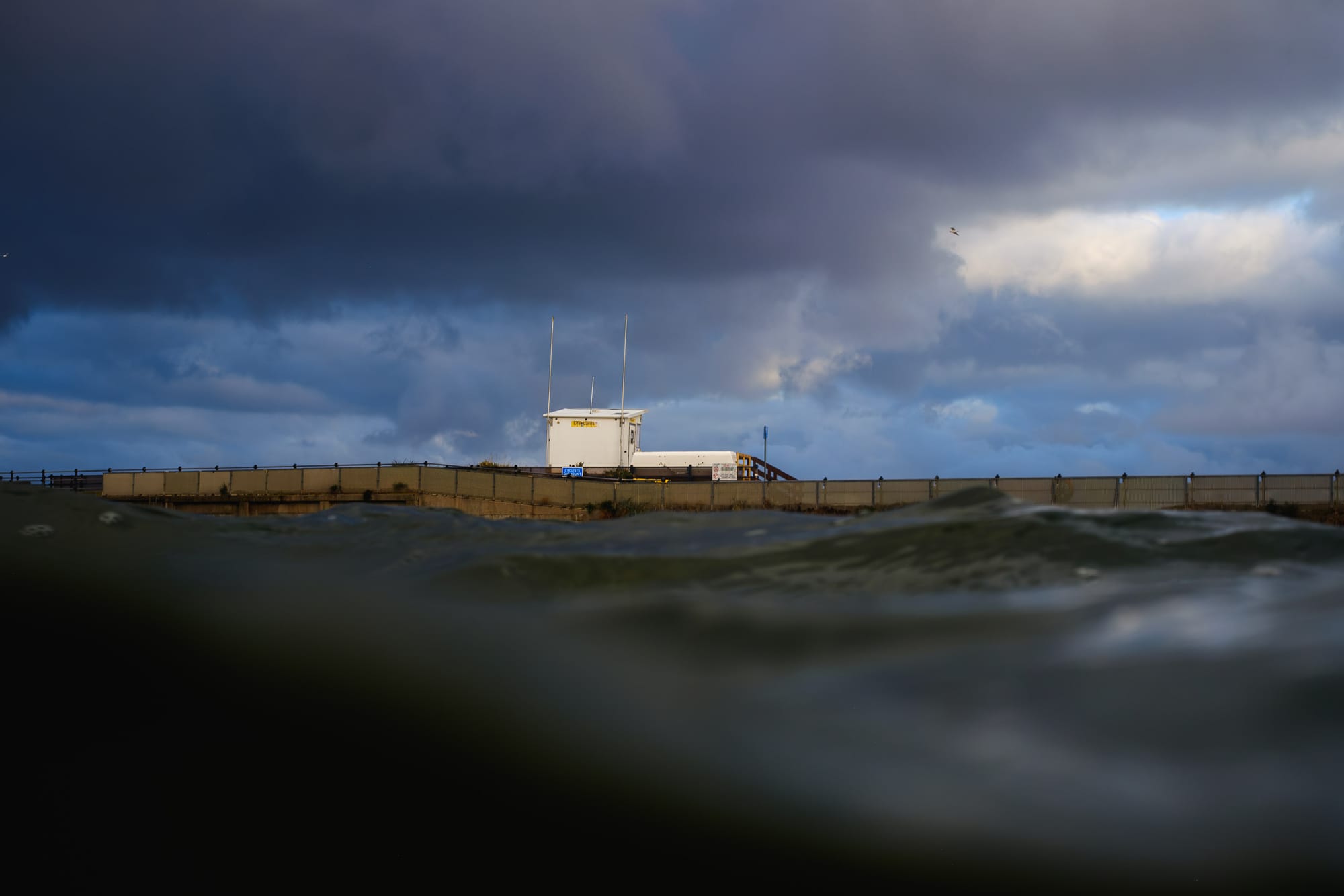 A lifeguard tower stands on a pier, with turbulent waves partially obscuring the view from the water's surface. Dark, moody clouds fill the sky, hinting at an impending storm, while a few scattered birds can be seen overhead. The scene conveys a sense of calm tension between the sea and the approaching weather.