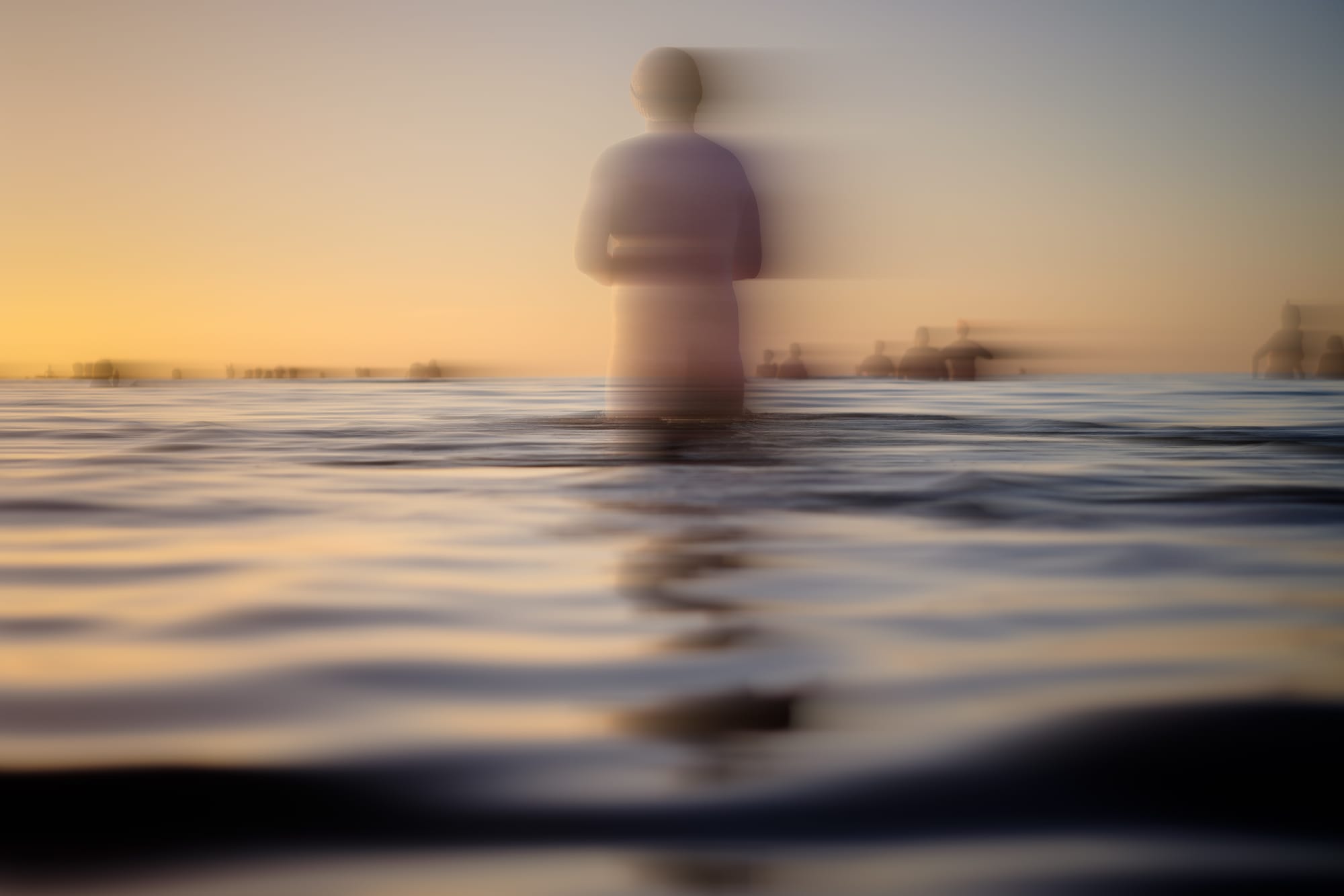 A naked person stands in the sea at sunrise as others splash about in the distance.