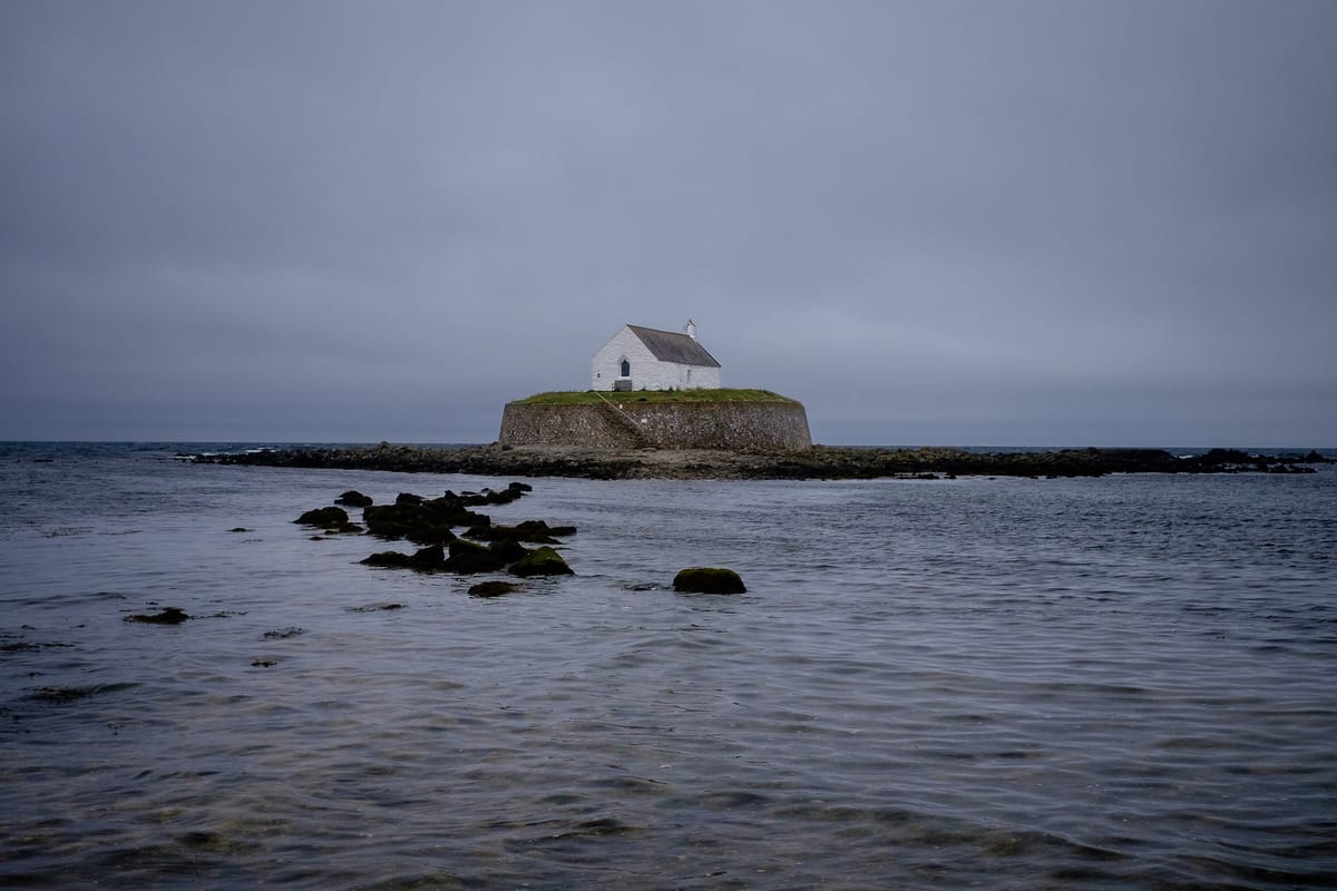 A small white house stands on a grassy island surrounded by shallow waters under a cloudy, gray sky. 