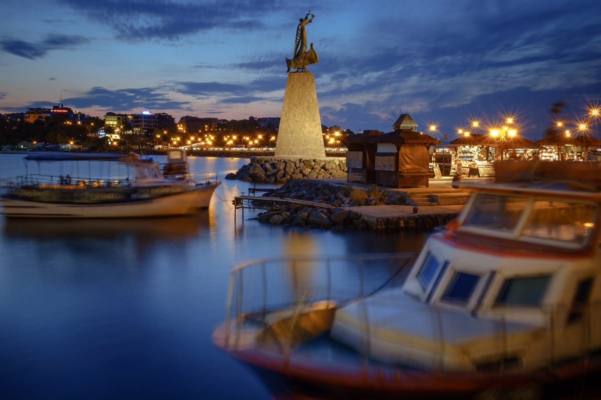 A serene evening scene by the water, featuring a prominent monument of a figure holding a sail, standing atop a stone pedestal. 