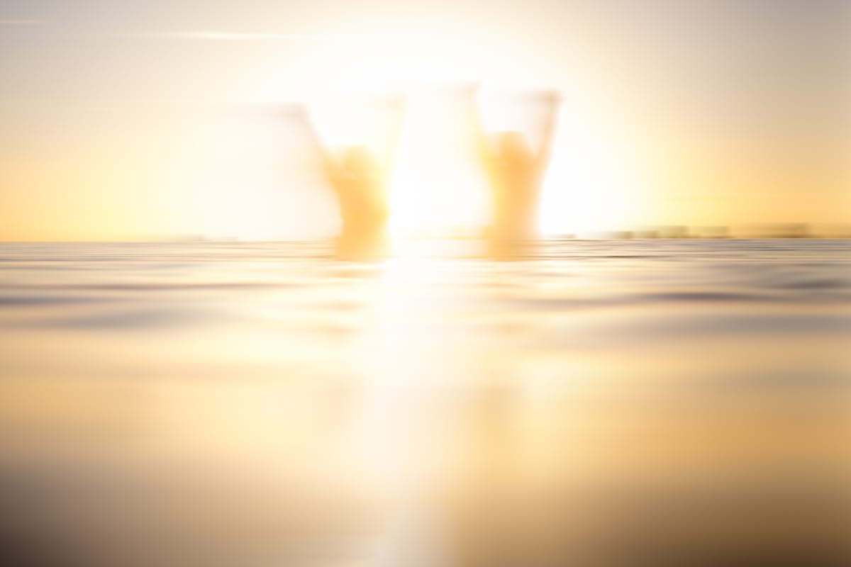 Two people hold their hands up in the air while standing in the sea at sunrise.