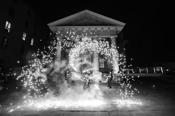 Performers dance with pyrotechnic devices. At the front a woman spins a stick spraying fireworks.