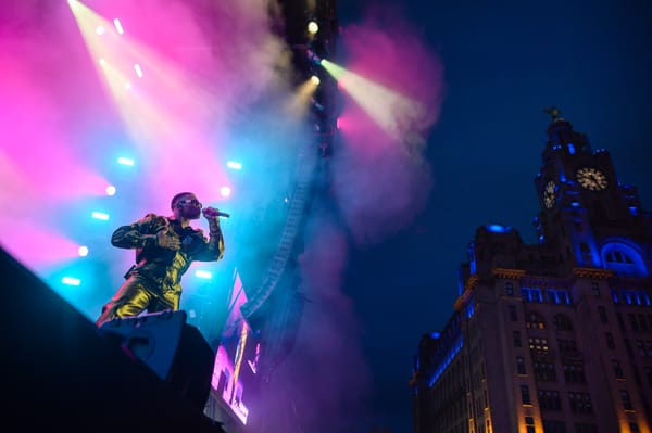 A singer lit by pink and blue lights looks up to the Liver Building lit up in blue and yellow.