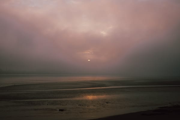 A misty landscape featuring a faint sun rising. The scene is shrouded in fog.