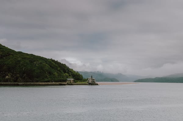 A photo of Barmouth, Wales, featuring a body of water, a shoreline with lush green vegetation, and a building on the edge of the water under an overcast sky.