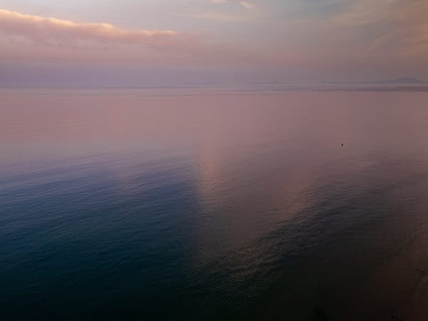 A serene coastal scene at dusk, featuring a vast expanse of water reflecting the soft hues of the sky. The water transitions from dark blue to a gentle pink, mirroring the clouds above.