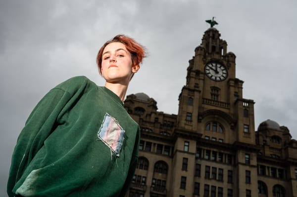 A person with red hair and a nose ring stands in front of the Royal Liver Building in Liverpool, England. The person is wearing a green sweatshirt. The sky is overcast.