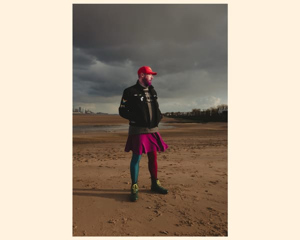 A person stands on a sandy beach under a dramatic sky, wearing a black jacket. They sport a vibrant purple skirt and colorful tights, complemented by bold green shoes.