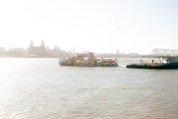 A colorful ferry boat sails on the River Mersey, with the Liverpool skyline in the background.