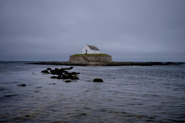 A small white house stands on a grassy island surrounded by shallow waters under a cloudy, gray sky. 