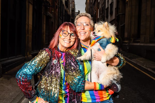 Two female presenting people in sparkly rainbow coats holding up a dog and smiling to camera.