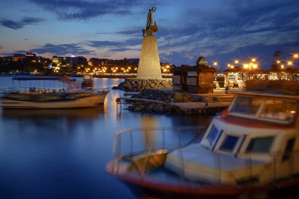 A serene evening scene by the water, featuring a prominent monument of a figure holding a sail, standing atop a stone pedestal. 