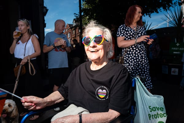 An older lady in a wheelchair wears rainbow heart sunglasses and smiles for the camera.