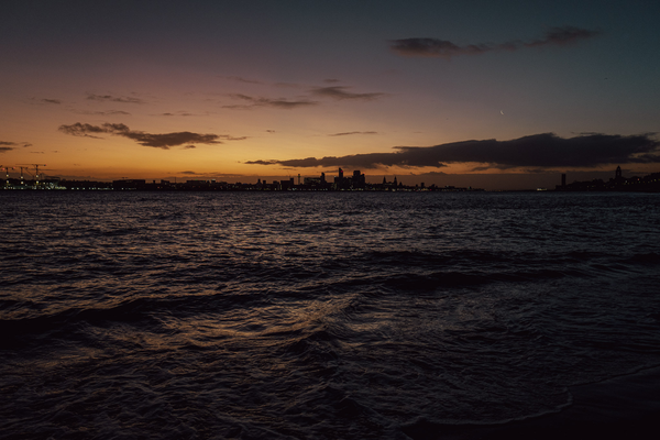 Dusk on the banks of the River Mersey. A new moon rises and there's a slight orange glow on the horizon behind the city of Li