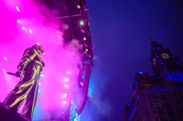 A member of Tvorchi band stands on the edge of a stage and looks up to the Liverbuilding lit up at dusk.