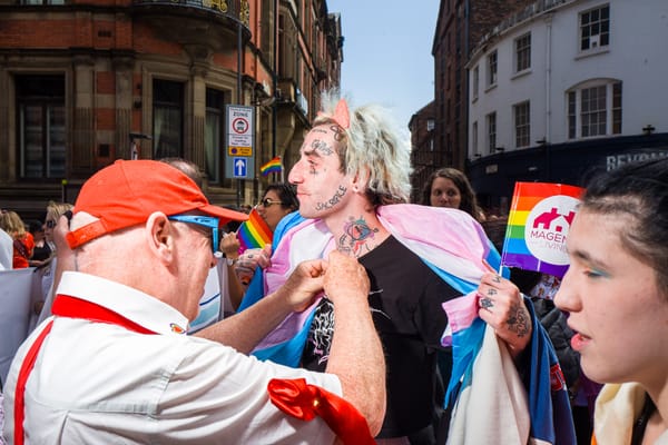 A person wearing a trans flag has a badge put on their t-shirt in the middle of a march.