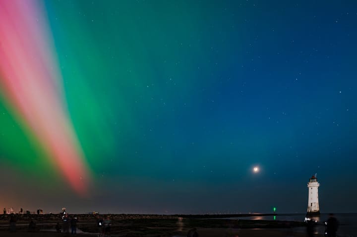 Red and green bands of light shimmer on the edge of the nights sky near a lighthouse.
