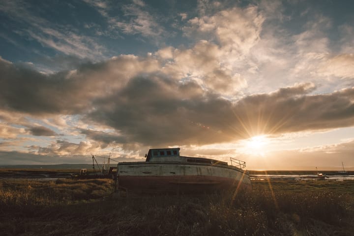 Sun sets behind an old worn out boat resting on a grassy riverbed.