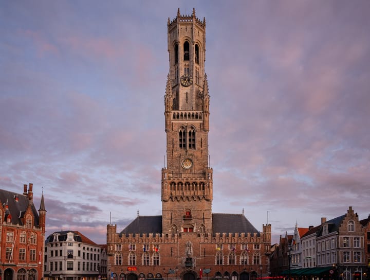 A tall clock tower extends high into the sky in the middle of Bruges at sunset.