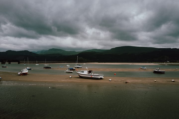 Lots of little boats rest on a river bed after the tide has gone out. Above them is a moody sky.
