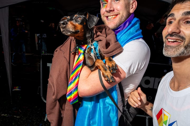 A white man holds up their little dog who is wearing a rainbow tie.