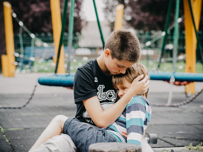 An older brother embraces his distressed sibling to comfort them while in a park.