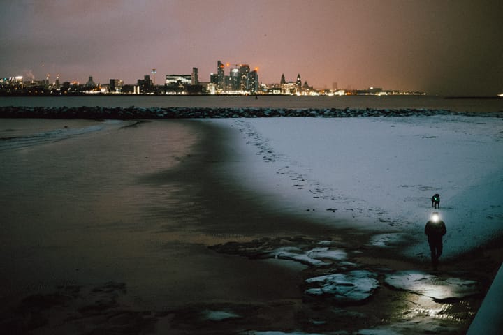 A person walking along a snow-covered beach at night, holding a flashlight.