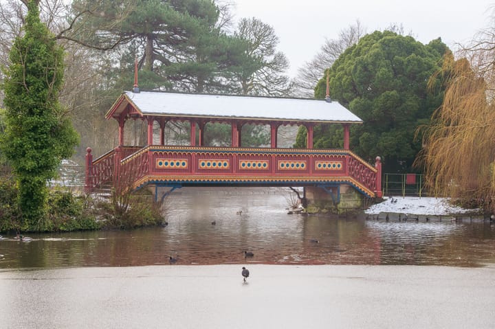 A decorative wooden bridge with a snow-covered roof spans across a body of water. 