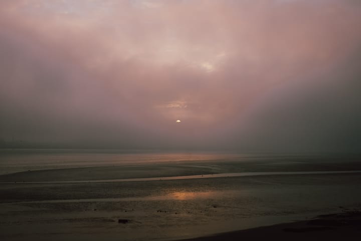 A misty landscape featuring a faint sun rising. The scene is shrouded in fog.