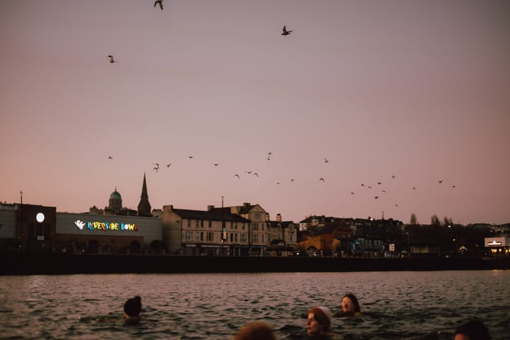 A scene depicting people swimming in a body of water. Birds are flying overhead against a twilight sky.