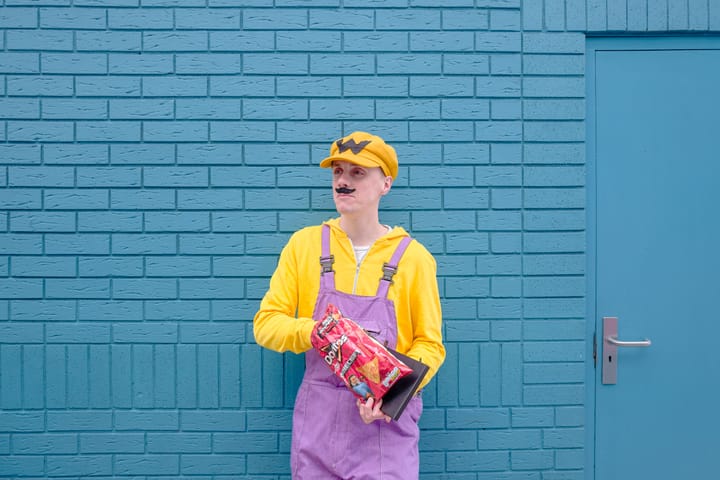 A person dressed in Wario cosplay stands in front of a blue brick wall, holding a bag of Doritos.