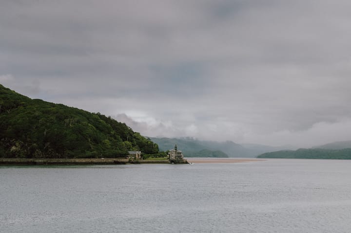 A photo of Barmouth, Wales, featuring a body of water, a shoreline with lush green vegetation, and a building on the edge of the water under an overcast sky.