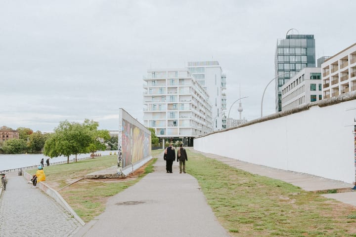 A segment of the Berlin Wall, with graffiti art, stands along a paved path. Two people walk along the path, with a body of water and modern buildings in the background.