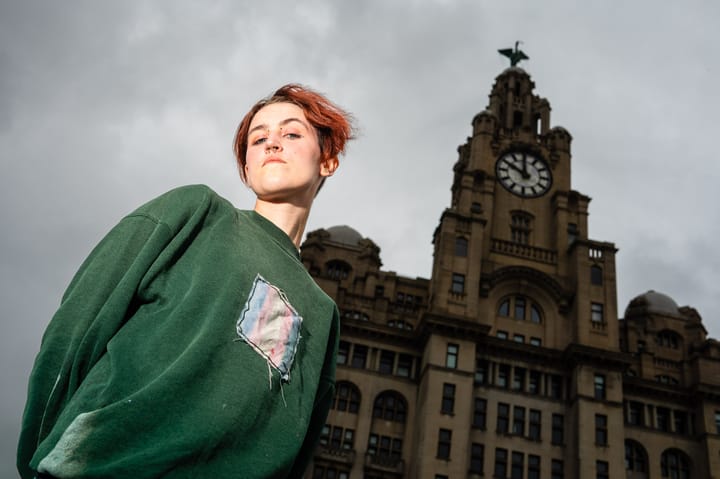 A person with red hair and a nose ring stands in front of the Royal Liver Building in Liverpool, England. The person is wearing a green sweatshirt. The sky is overcast.