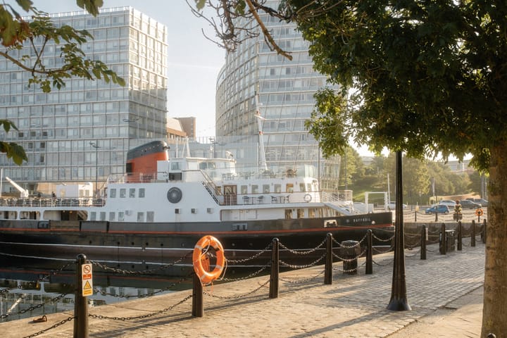 A view of the Liverpool waterfront at sunrise, featuring a boat, modern buildings, and a tree-lined promenade.