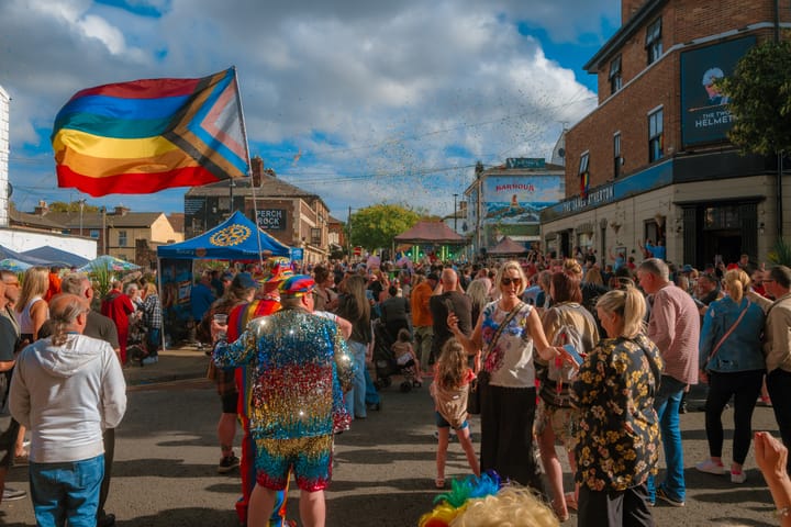 A vibrant street scene at Wirral Pride 2025 in New Brighton, with a large crowd of people, a rainbow flag, and confetti in the air.