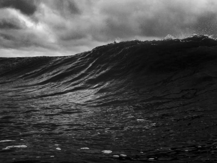 A dramatic black and white image of a large ocean wave, capturing its crest and the texture of the water.