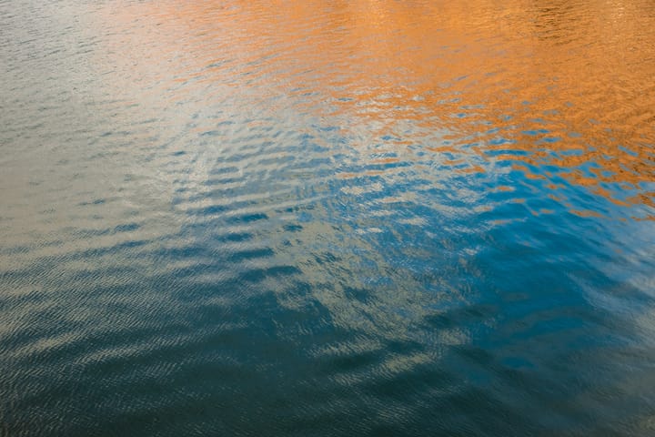 Abstract image of water surface with reflections in shades of blue, orange, and gray, with ripples and textures.